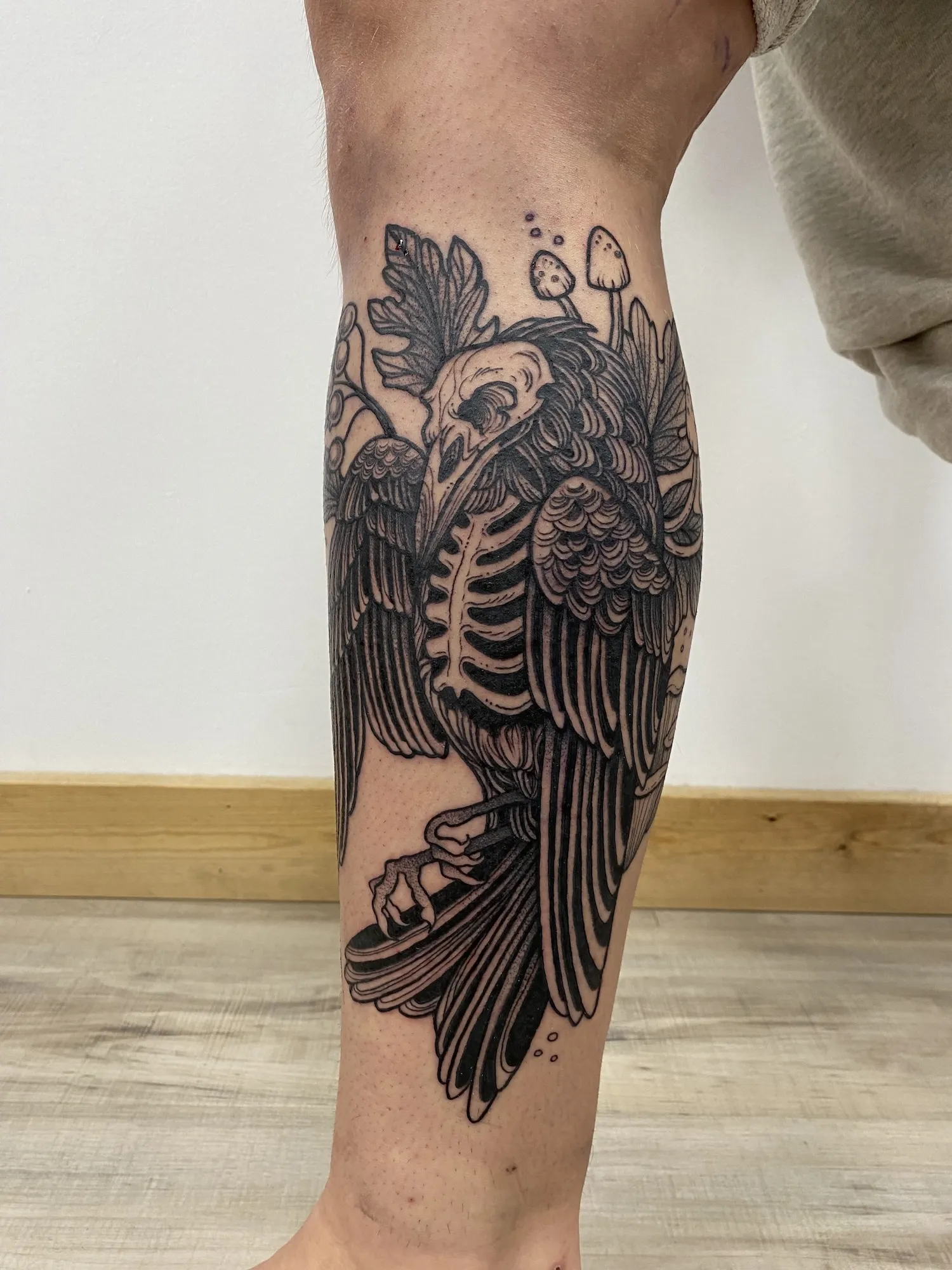 Blackwork upper arm tattoo of a skeletal crow with a skull head, leaves and mushrooms, by Alyssa at Vale of the Fae in Castlegar, BC