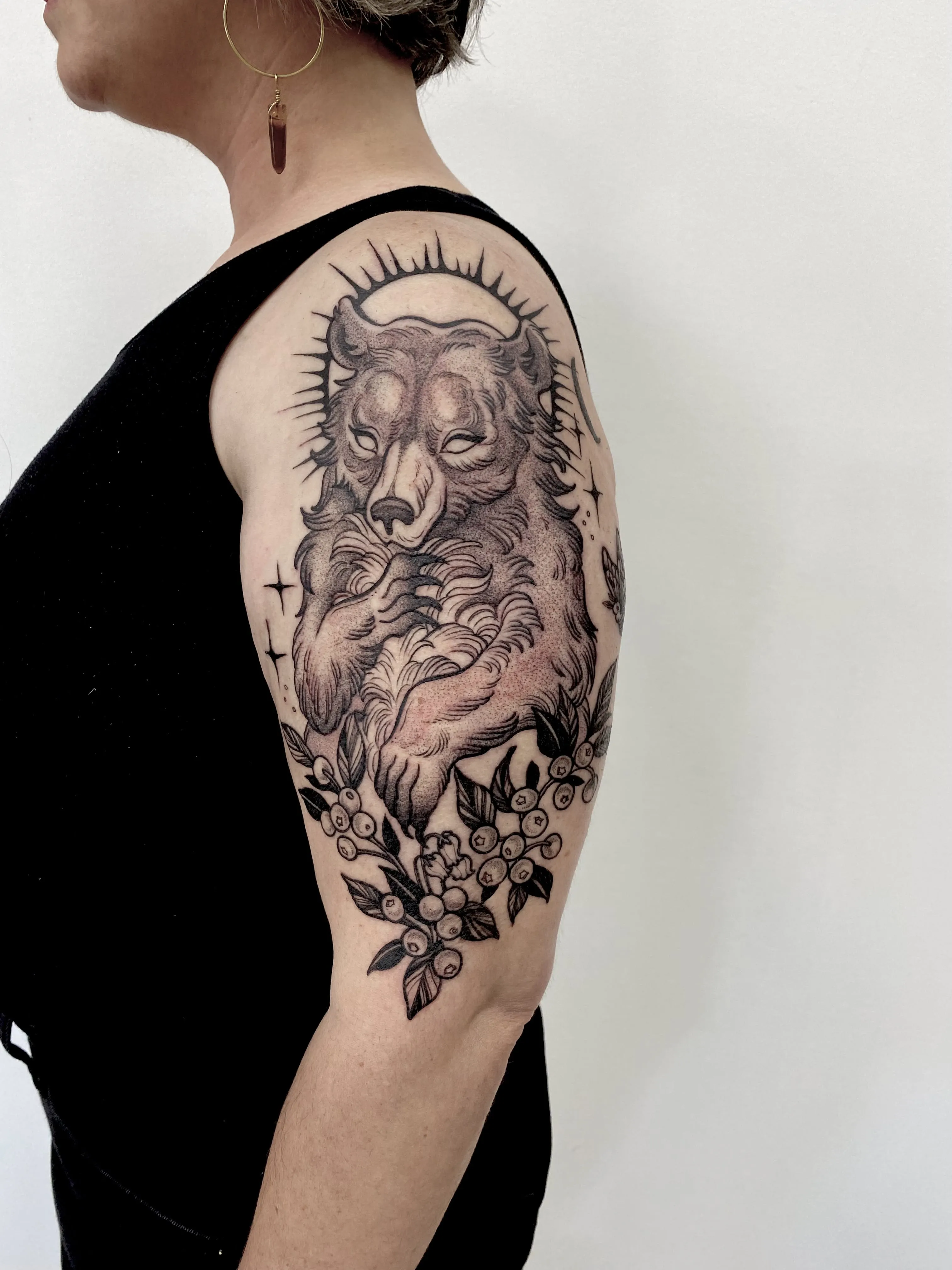 Black and grey haloed bear tattoo with stars and berries on the upper arm, by Alyssa at Vale of the Fae in Castlegar, BC