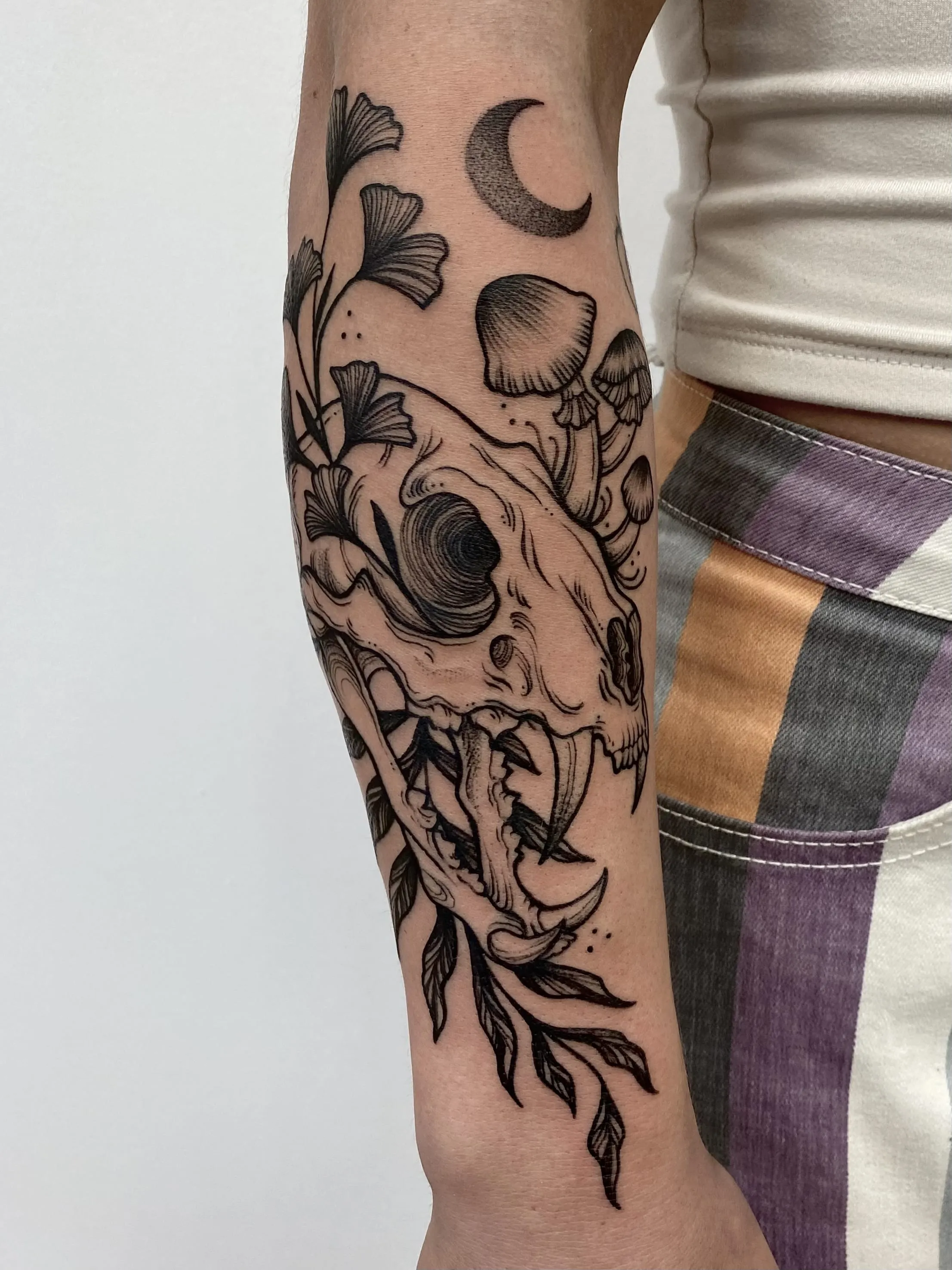Blackwork forearm tattoo of a fanged animal skull with leaves, mushrooms and a crescent moon, by Alyssa at Vale of the Fae in Castlegar, BC