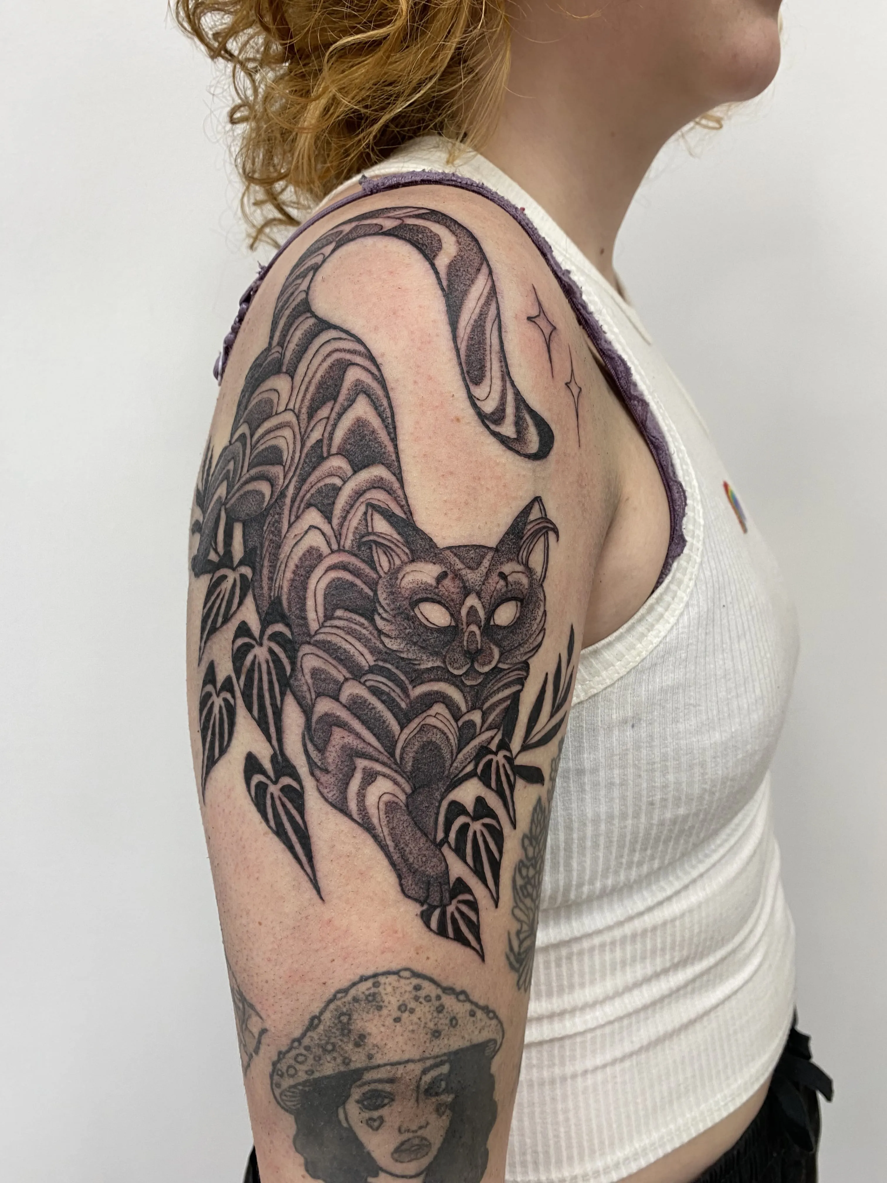 Blackwork upper arm tattoo of a stylized cat with swirling patterns stretching among leaves, by Alyssa at Vale of the Fae in Castlegar, BC