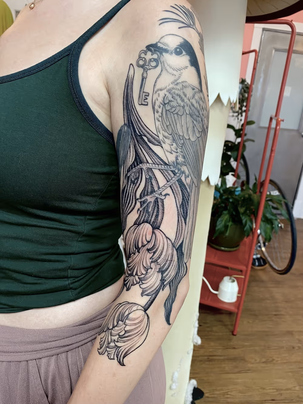 Blackwork upper arm tattoo of a bird holding an ornate key perched on tulips, by Alyssa at Vale of the Fae in Castlegar, BC