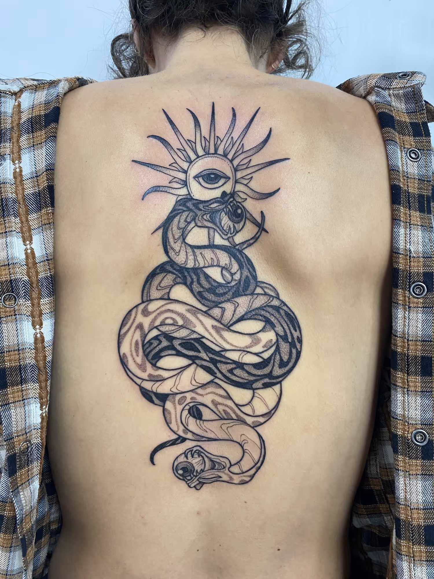 Blackwork back tattoo of a coiled patterned snake beneath a sun with an eye, by Alyssa at Vale of the Fae in Castlegar, BC