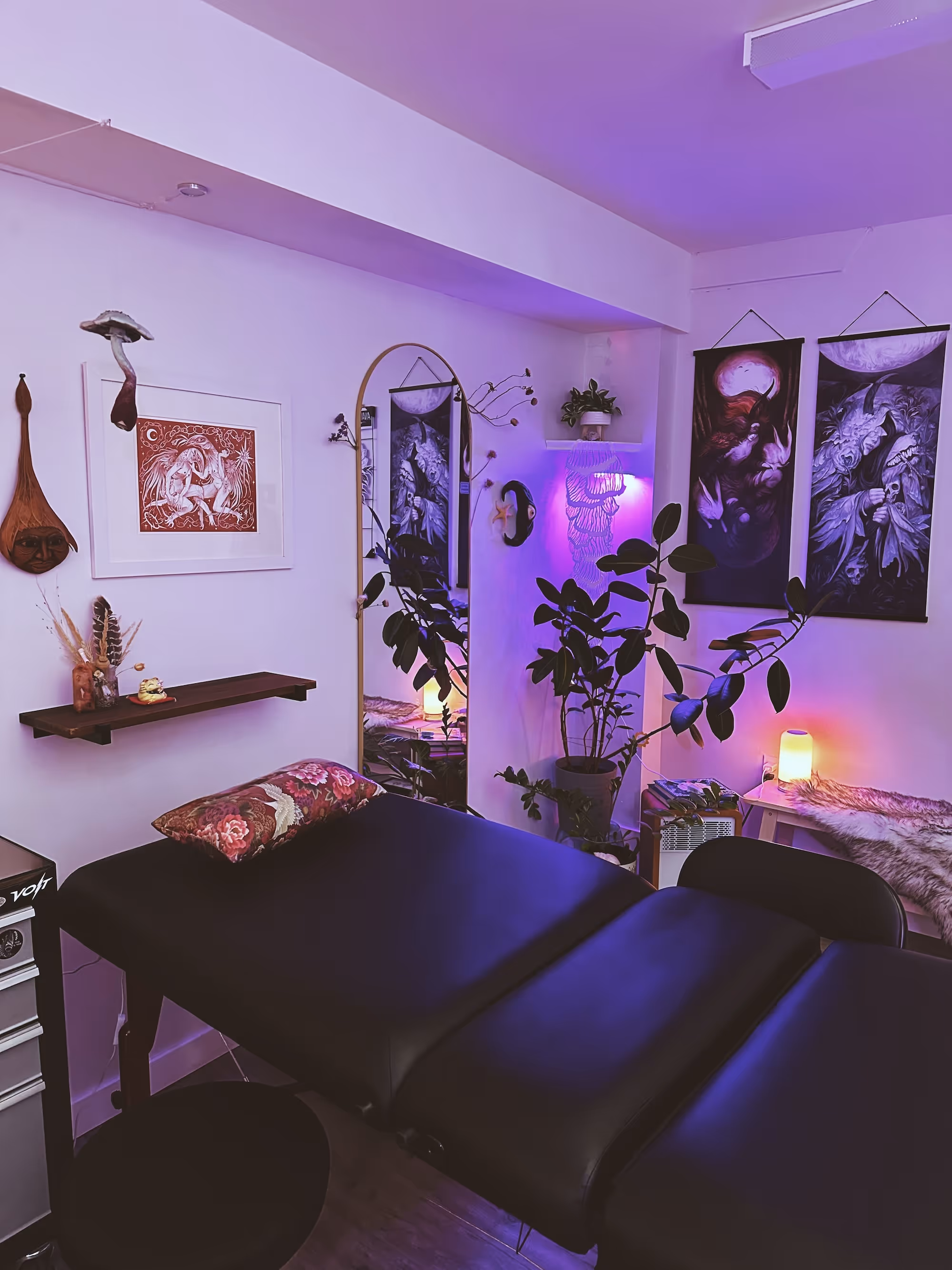 Interior of a cozy, dimly lit room with a black massage table, floral pillow, hanging art, a tall mirror, potted plant, and purple ambient lighting.