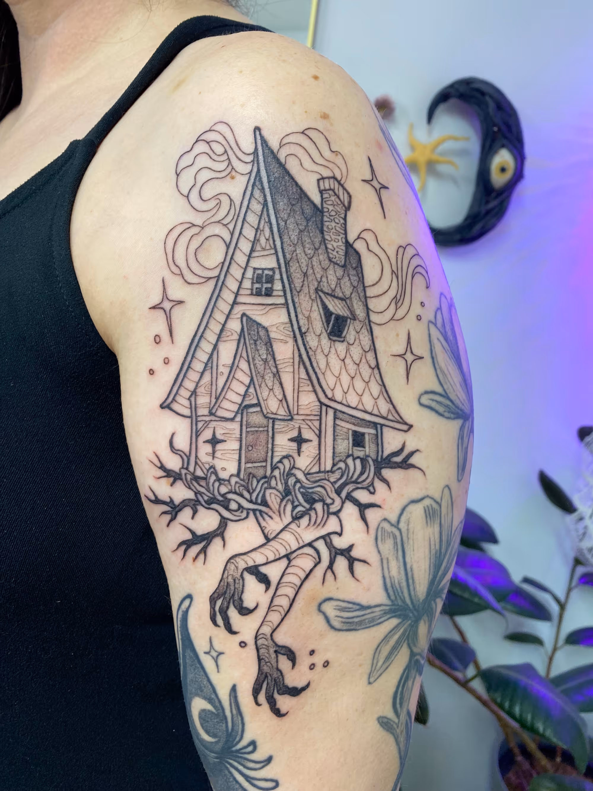 Blackwork upper arm tattoo of a witch house with clawed roots, stars and smoke, by Alyssa at Vale of the Fae in Castlegar, BC