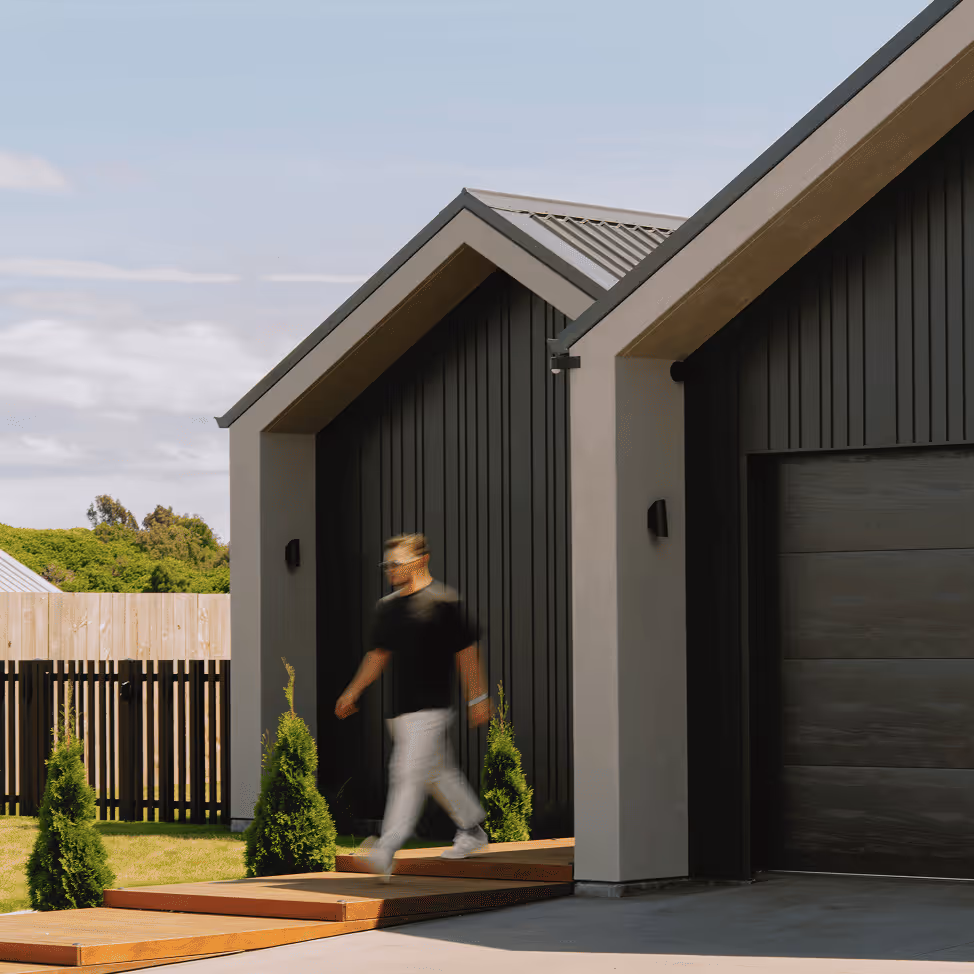 Person walking on wooden pathway beside modern black and gray house with a garage and small pine trees.