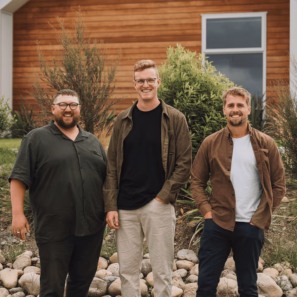 Three smiling men standing outdoors in front of rocks, greenery, and a wooden building with a window.