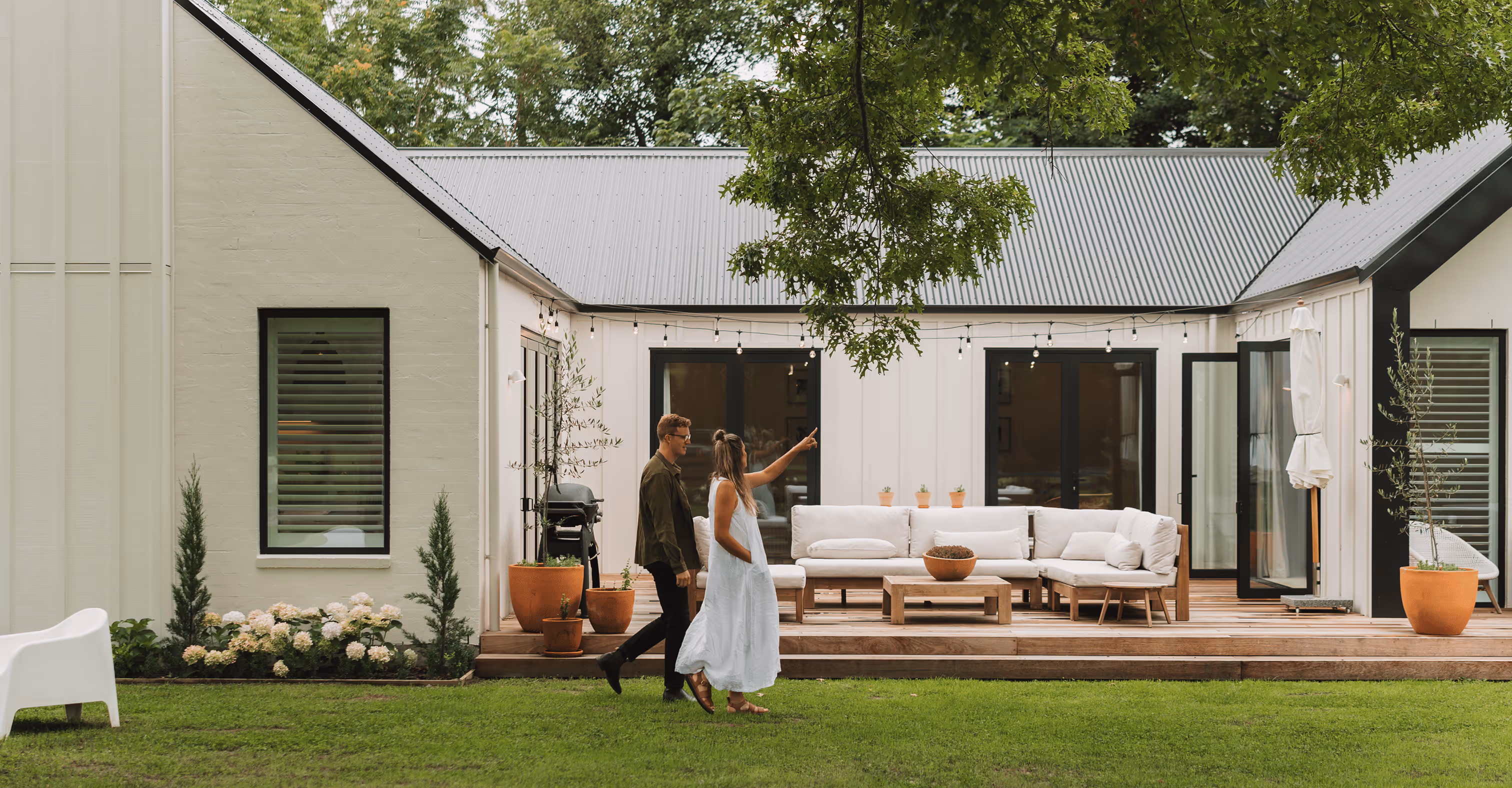 Couple walking on grass pointing towards a modern white house with a wooden deck and outdoor seating.