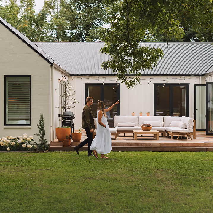 A couple walking on a grassy lawn in front of a modern white house with a patio featuring a white sectional sofa and wooden coffee table.