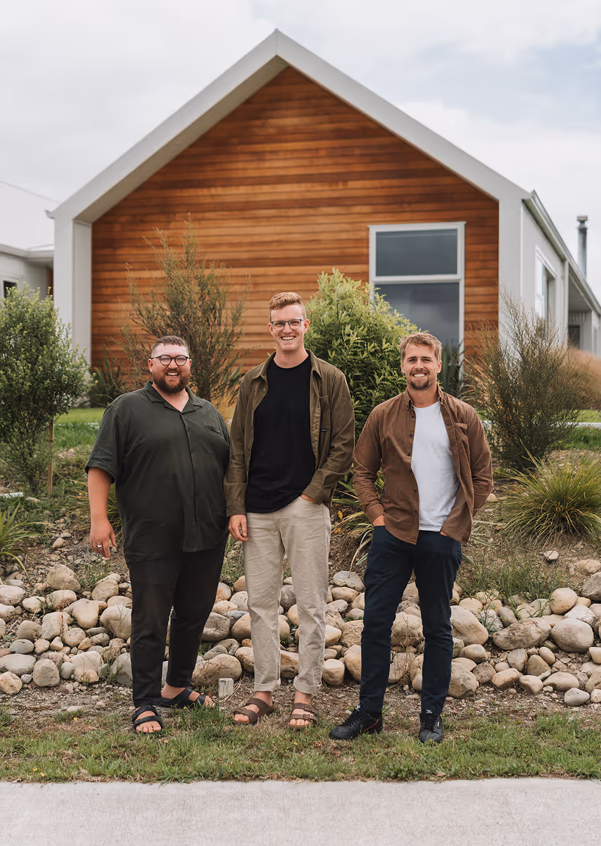 Three smiling men standing side by side on grass in front of a modern house with wooden siding and white trim.