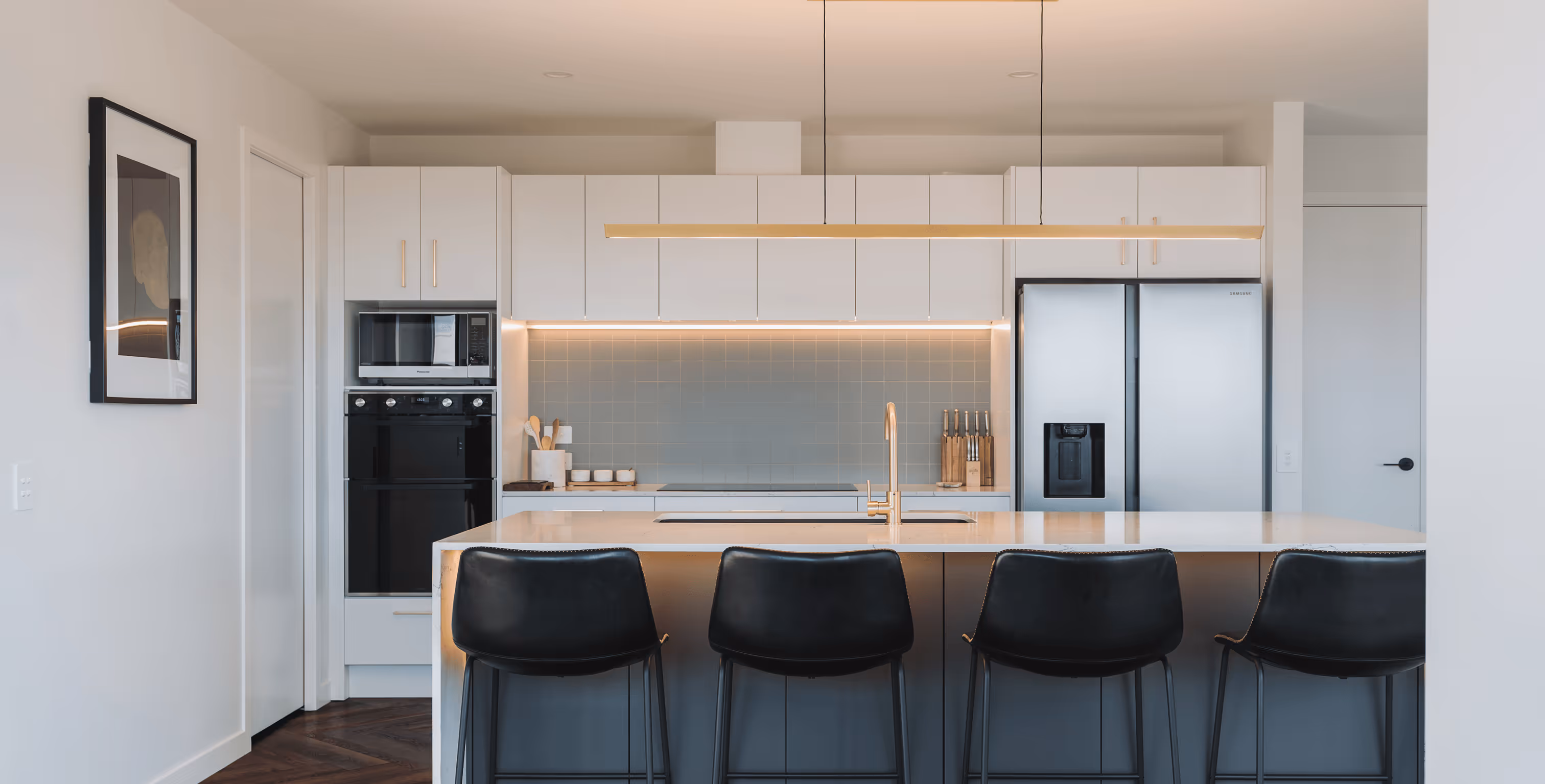 Modern kitchen with white cabinets, light blue tiled backsplash, stainless steel fridge, built-in oven, and four black bar stools at a white island.