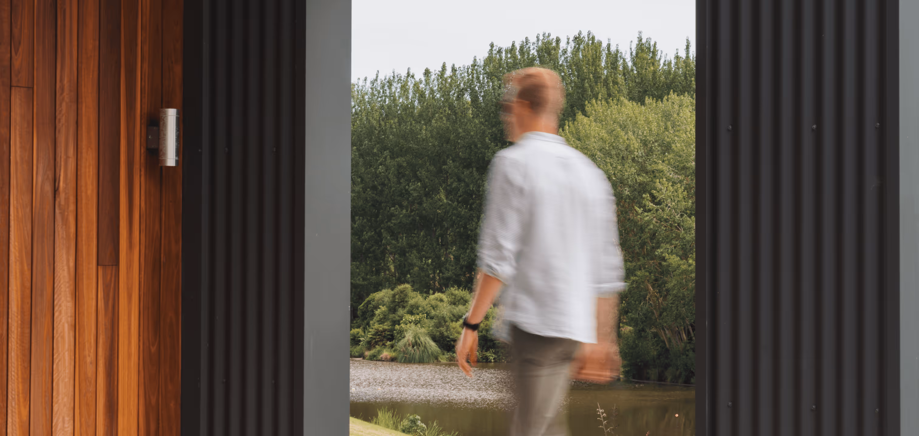 Blurred man walking outside near a wooden and metal doorway with green trees and a pond in the background.