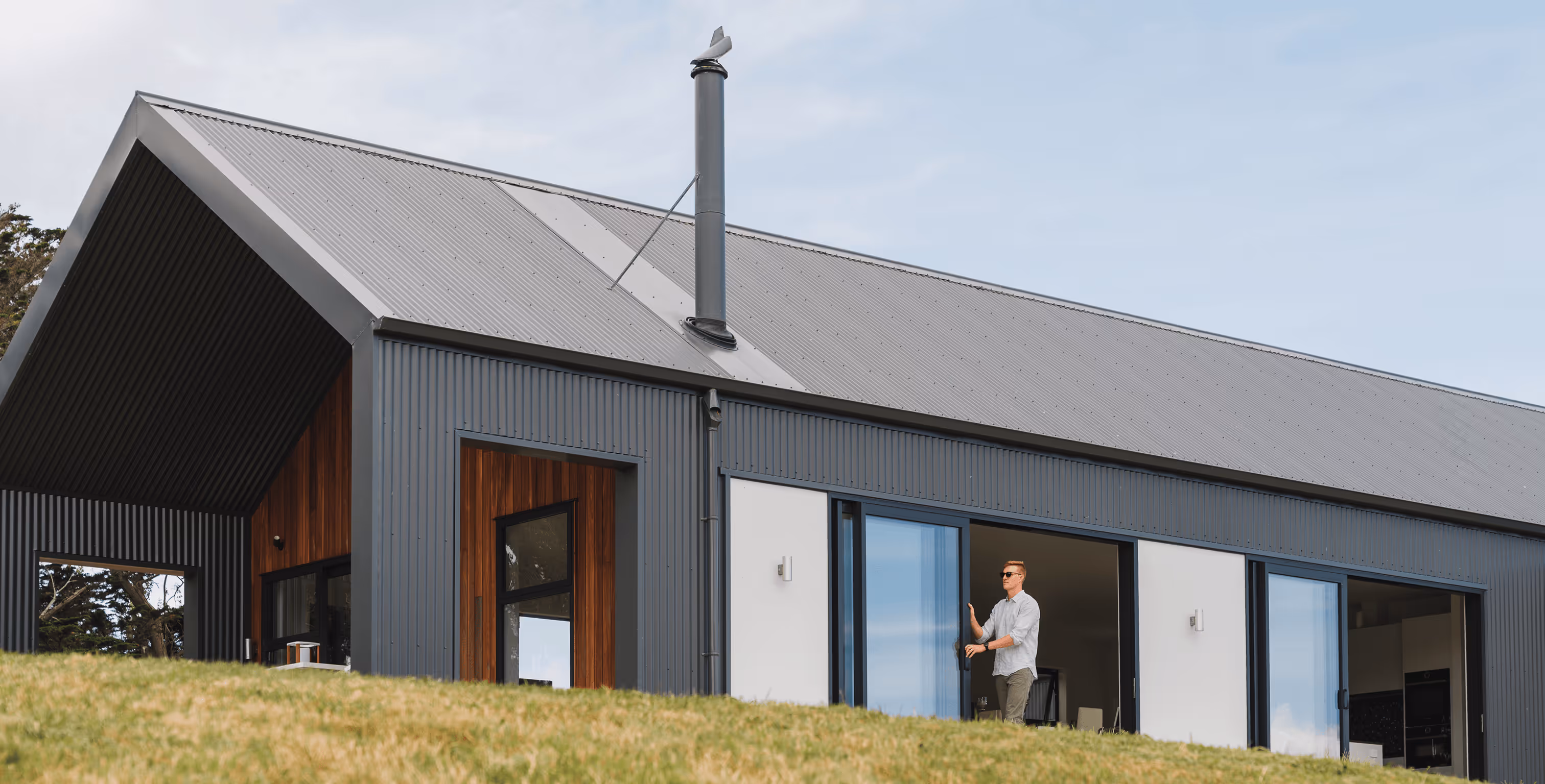Modern house with dark gray corrugated metal siding and roof, large glass doors, and a person standing in the doorway.