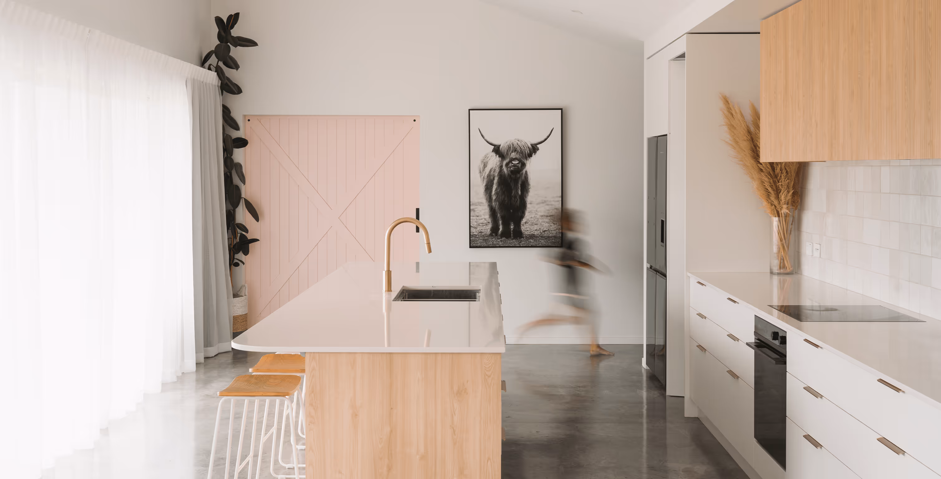 Modern kitchen with a wooden island, white countertop, gold faucet, stools, a black and white highland cow photo, and a blurred person walking.