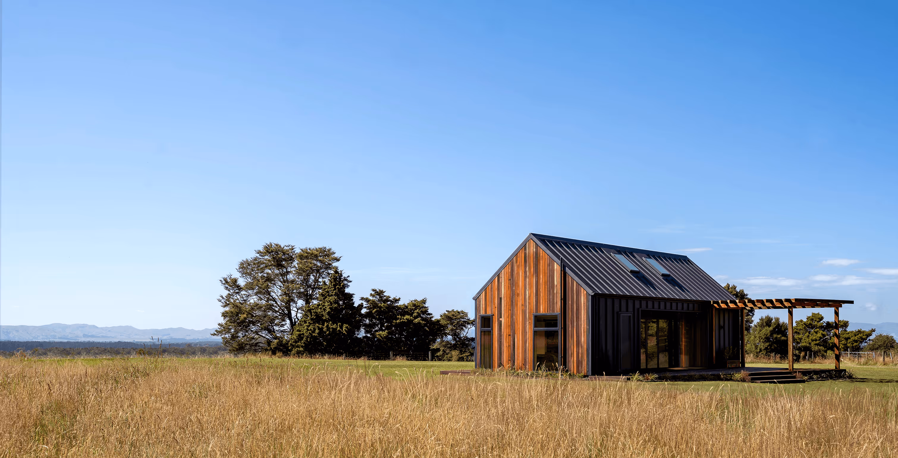 Modern small house with wood and black metal siding in a grassy field under clear blue sky.