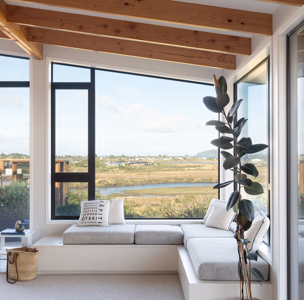 Modern corner seating area with gray cushions and windows offering a view of a river and open landscape under wooden ceiling beams.