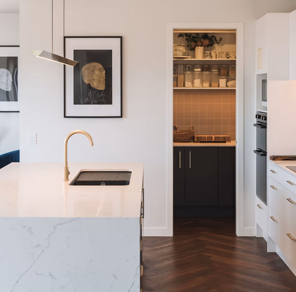 Modern kitchen with marble island, gold faucet, built-in ovens, and a pantry with glass jars and mugs.