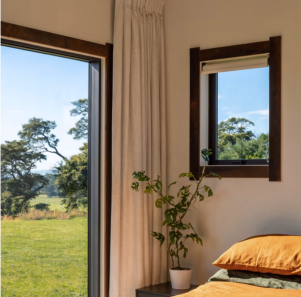 Indoor bedroom corner with a potted plant on a nightstand, a small window with a view of trees, and a large open door showing a green grassy field.