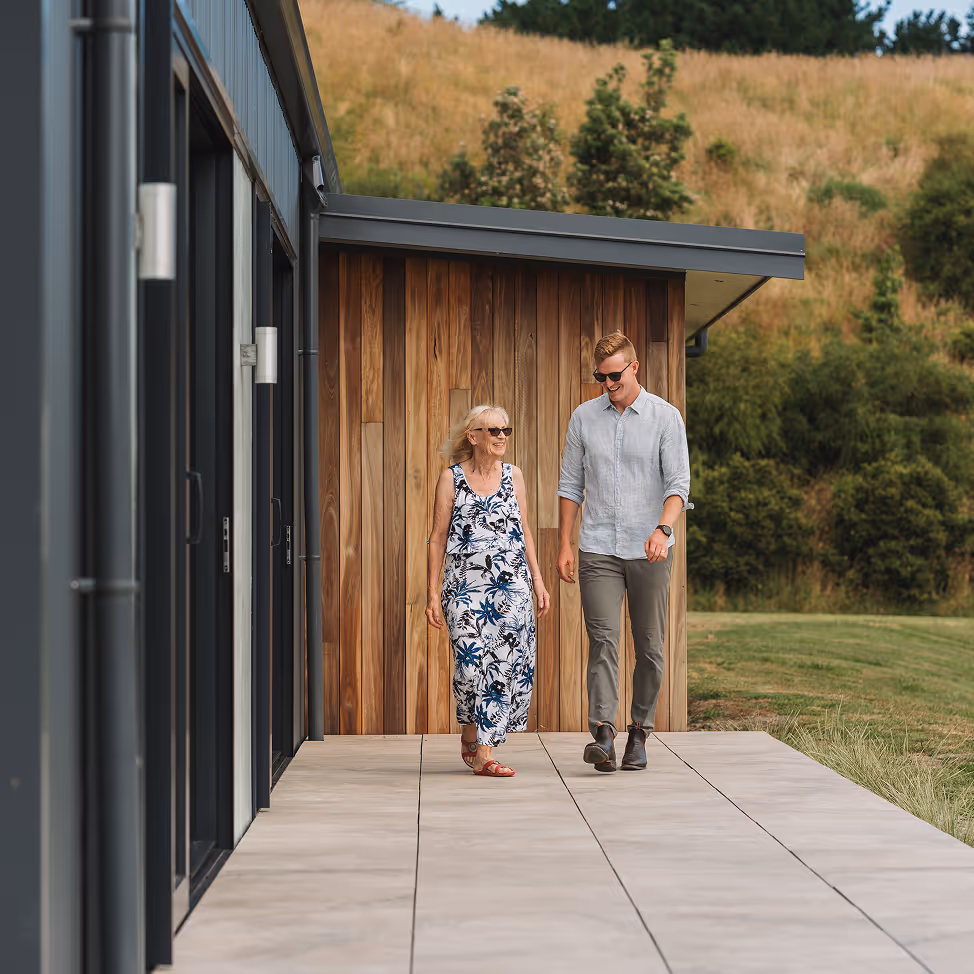 Elderly woman and young man walking and smiling on a tiled patio outside a modern building with wooden siding and grassy hillside in the background.