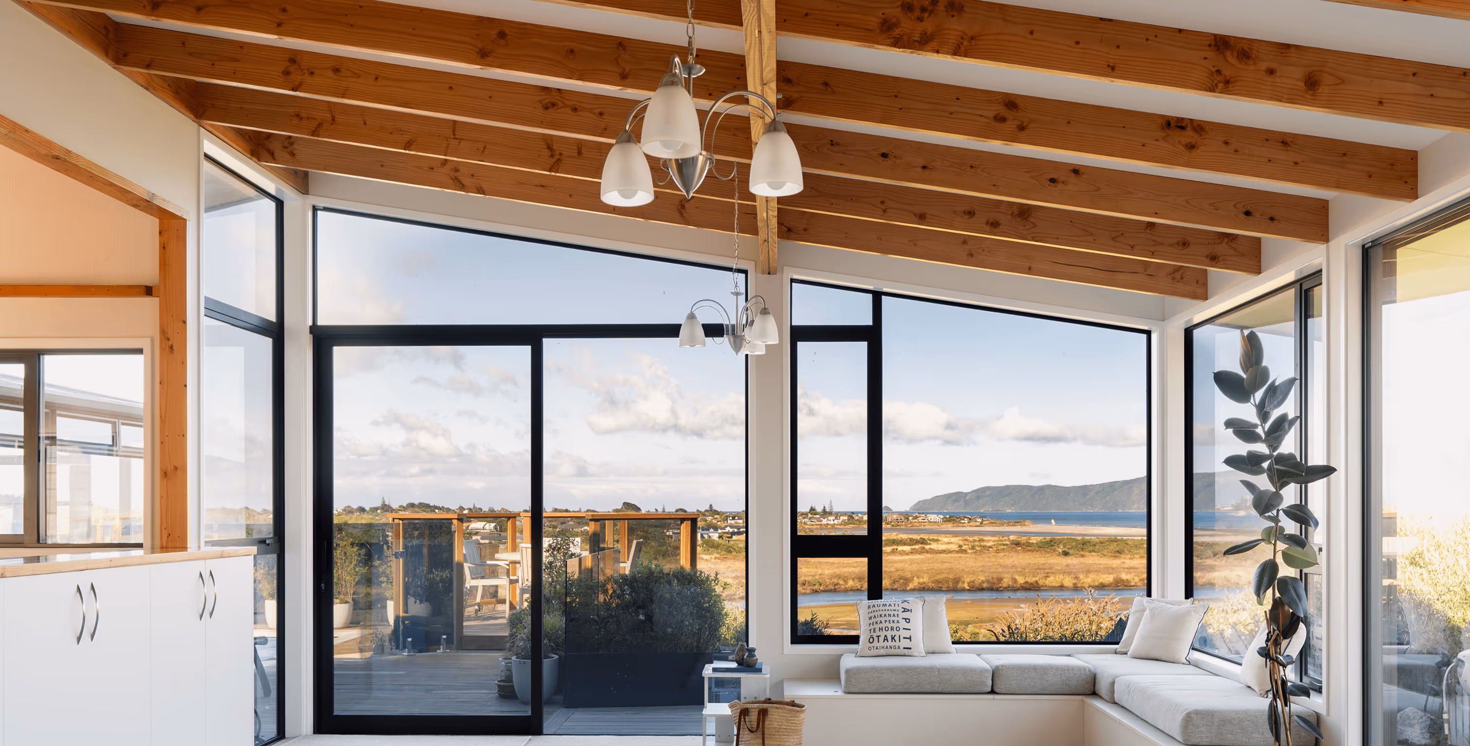 Living room with large windows, wood ceiling beams, white sectional sofa, and a scenic view of water and hills outside.