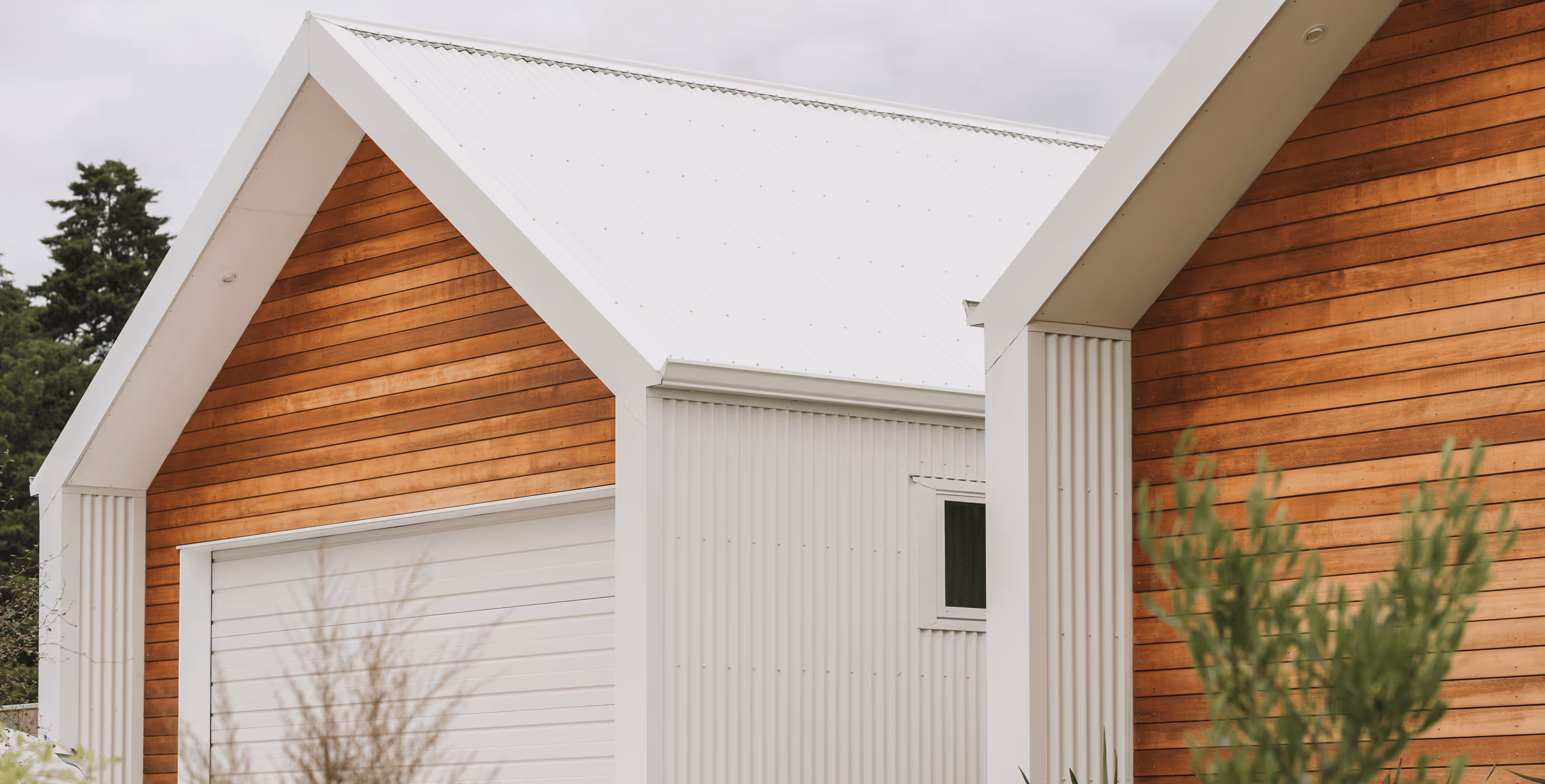Modern house exterior with white corrugated metal siding, wooden paneling, white garage door, and greenery in front.