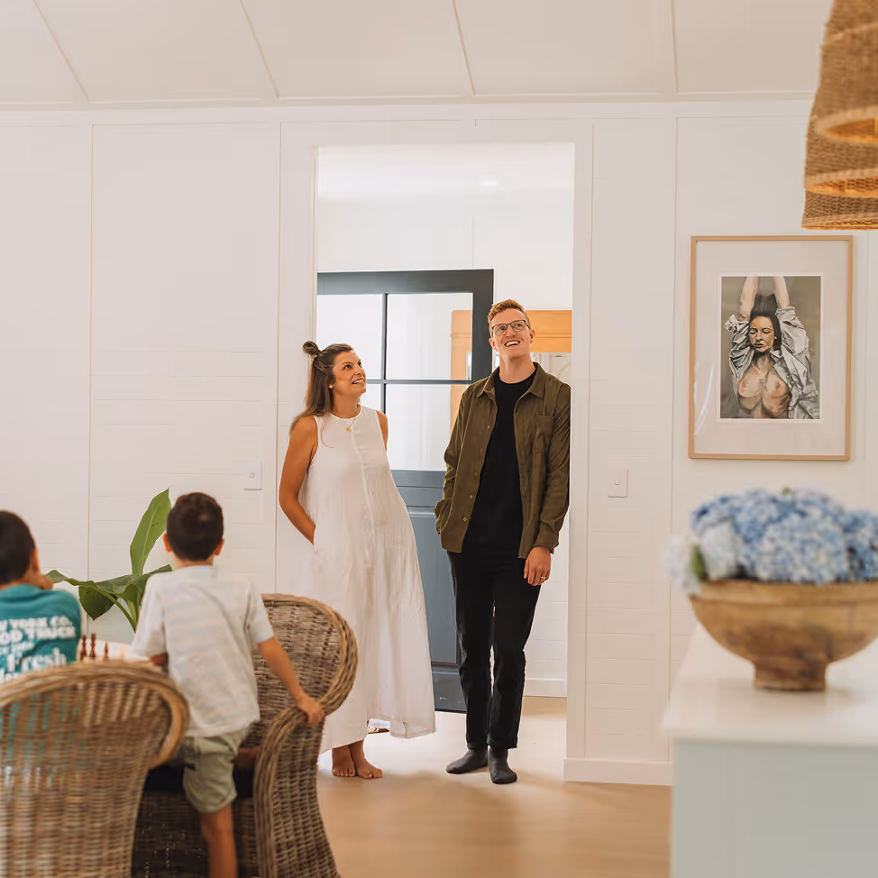 A smiling couple standing barefoot in a doorway looking at two children sitting at a wicker table inside a bright, modern home.