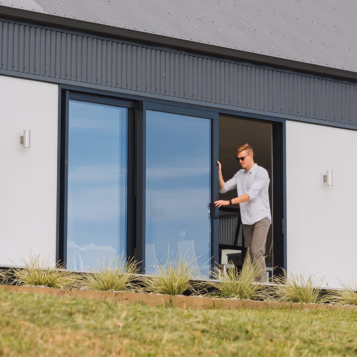 Man wearing sunglasses sliding open a large glass door of a modern house with a garden in front.