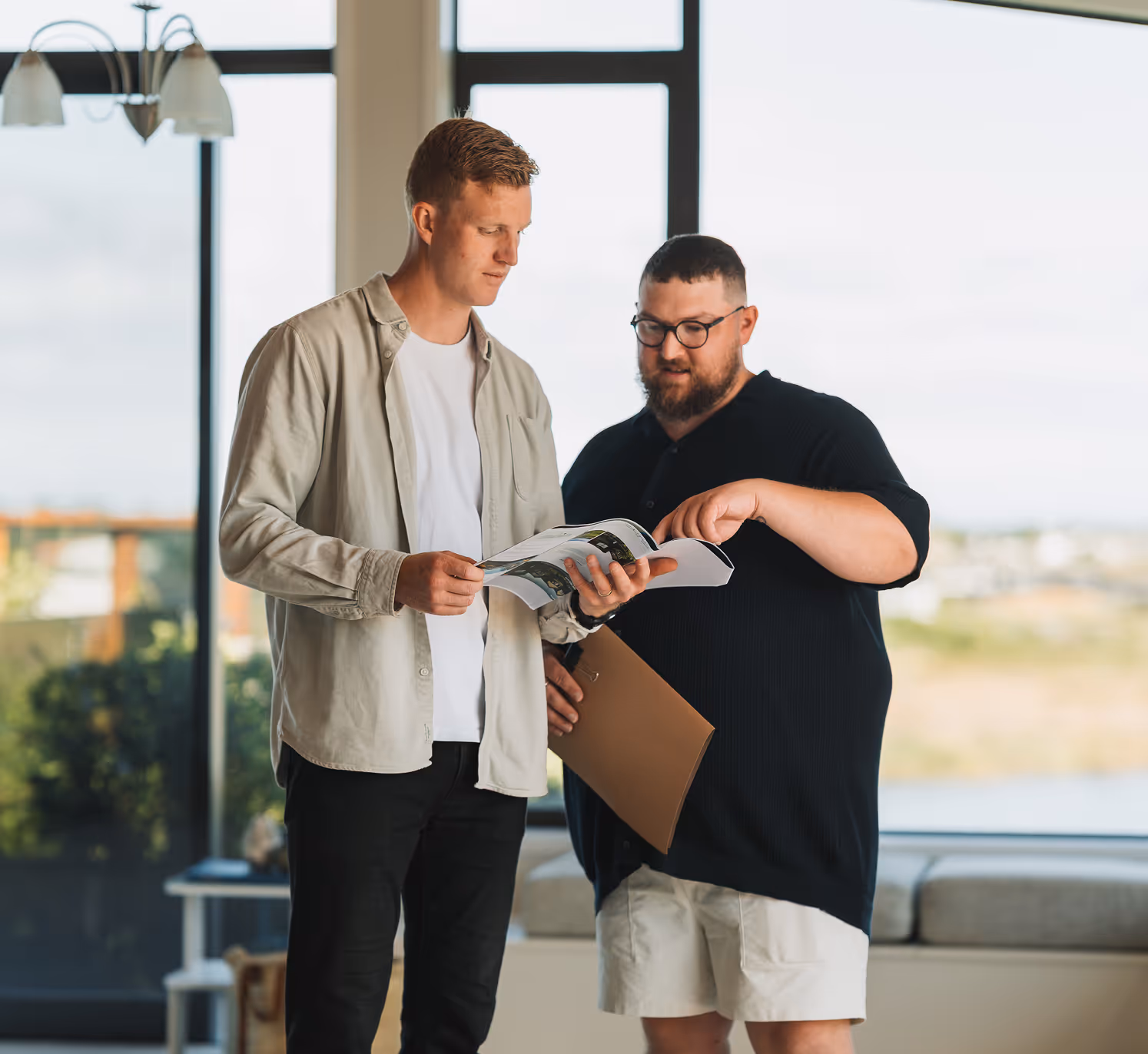 Two men standing indoors looking at and discussing an open magazine or booklet.
