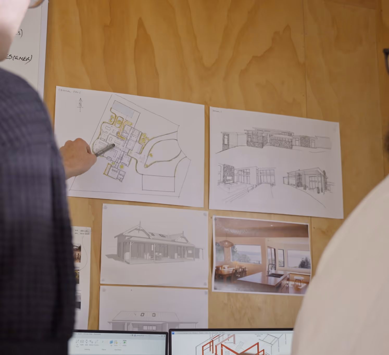 Two people reviewing architectural plans and sketches pinned on a wooden wall above computer screens.
