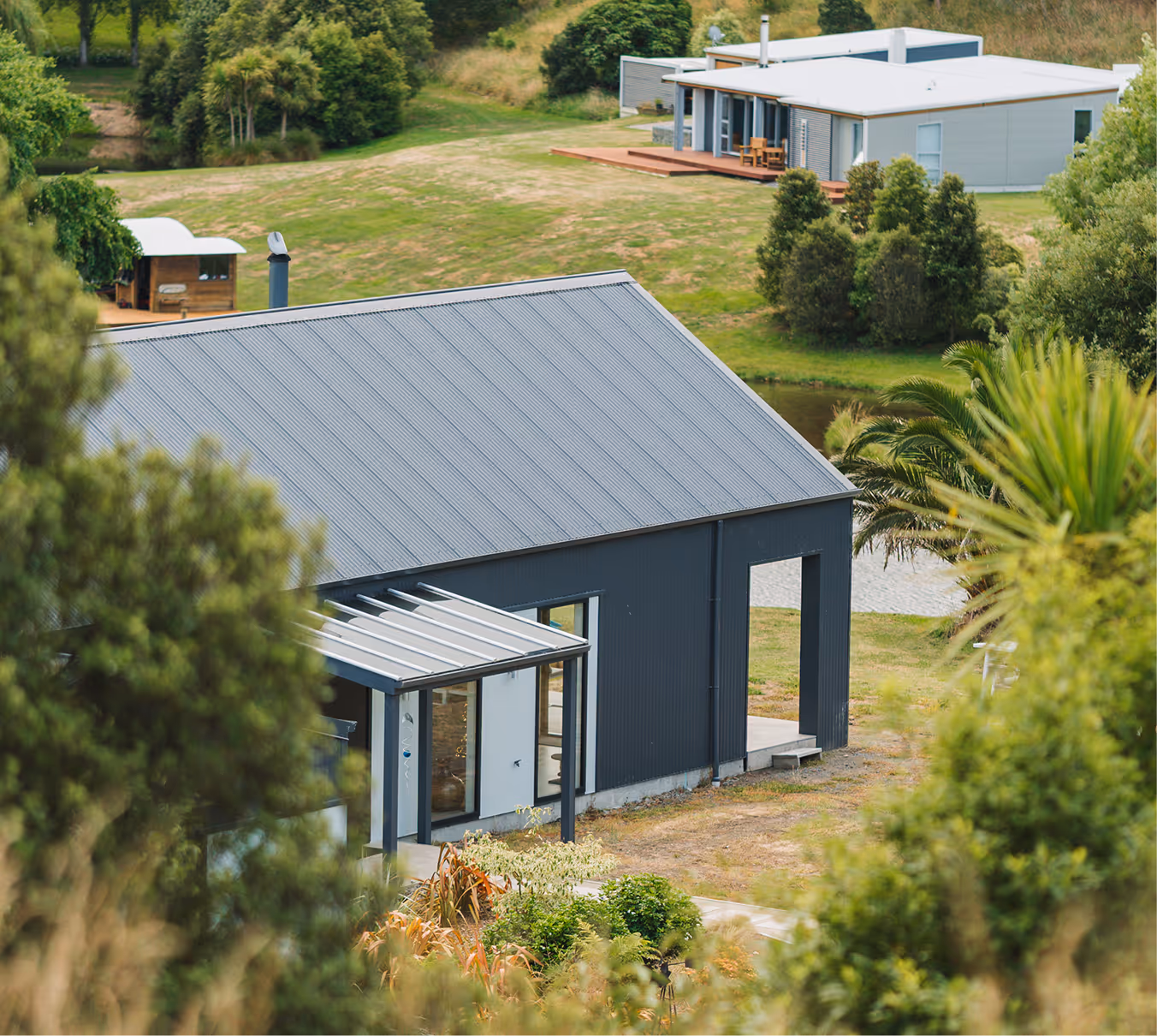 Modern dark gray house with a metal roof and pergola entrance, surrounded by green trees and grass, with another white modern house in the background.