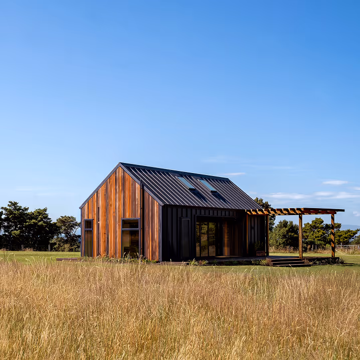 Modern small house with wooden siding and black metal roof in a grassy field under a clear blue sky.