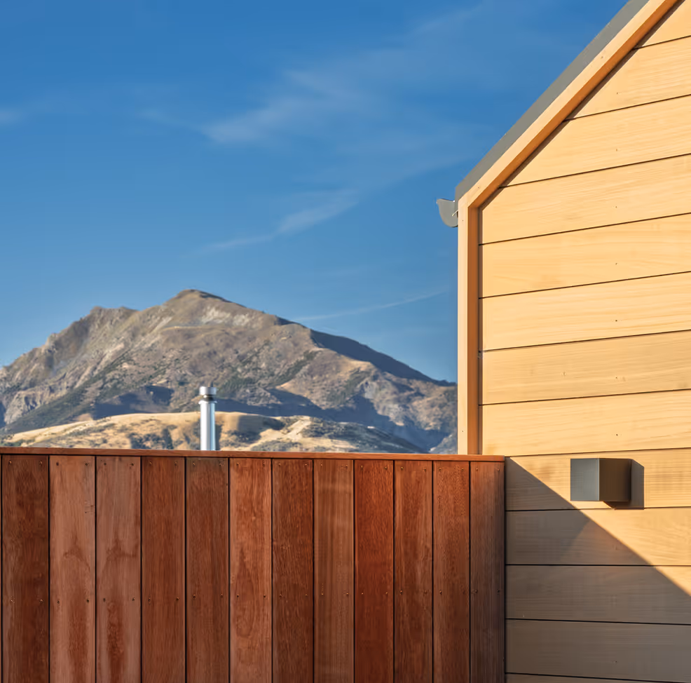 Mountain view behind a wooden fence and the corner of a house with wooden siding and a wall light.