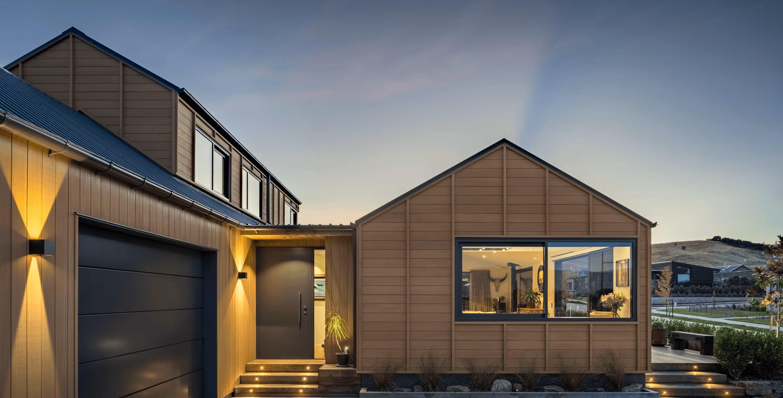 Modern two-story house with wooden exterior, black garage door, illuminated entrance, and large window showing interior decor at dusk.