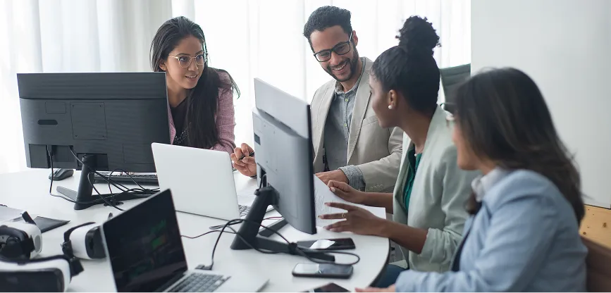 Four diverse professionals collaborate around a table with laptops, monitors, and tech devices in an office.