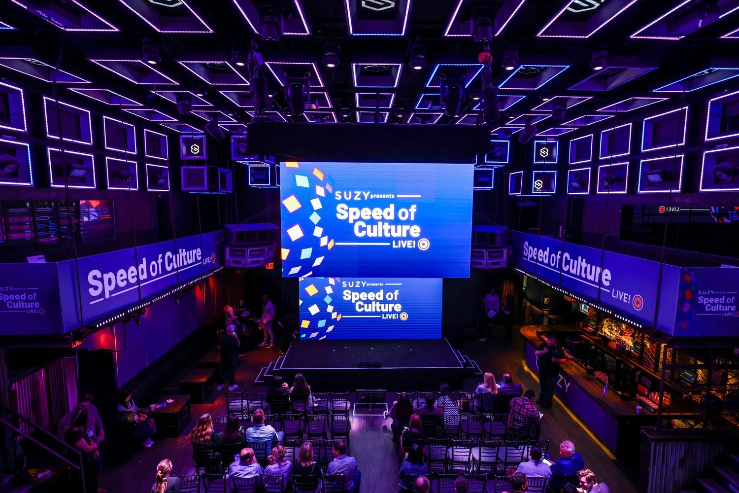 Audience seated in a dark venue with purple neon lights facing a stage with two large screens displaying 'SUZY presents Speed of Culture LIVE!'