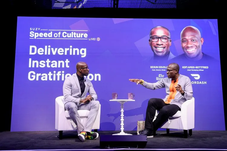 Two men seated on a stage engaged in a discussion with a purple backdrop displaying 'Speed of Culture LIVE! Delivering Instant Gratification' and photos of the speakers.