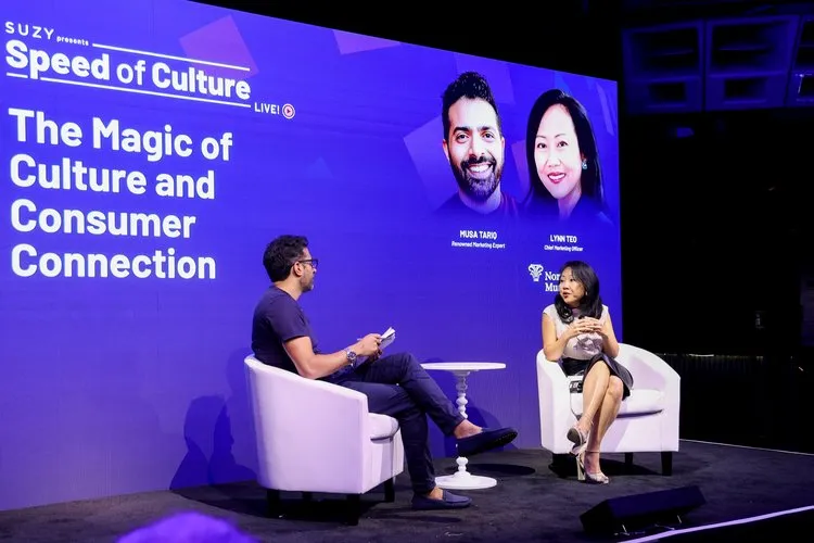 Two people seated on white chairs on stage in front of a large purple screen displaying 'Speed of Culture: The Magic of Culture and Consumer Connection' with photos and titles of Musa Tariq and Lynn Teo.
