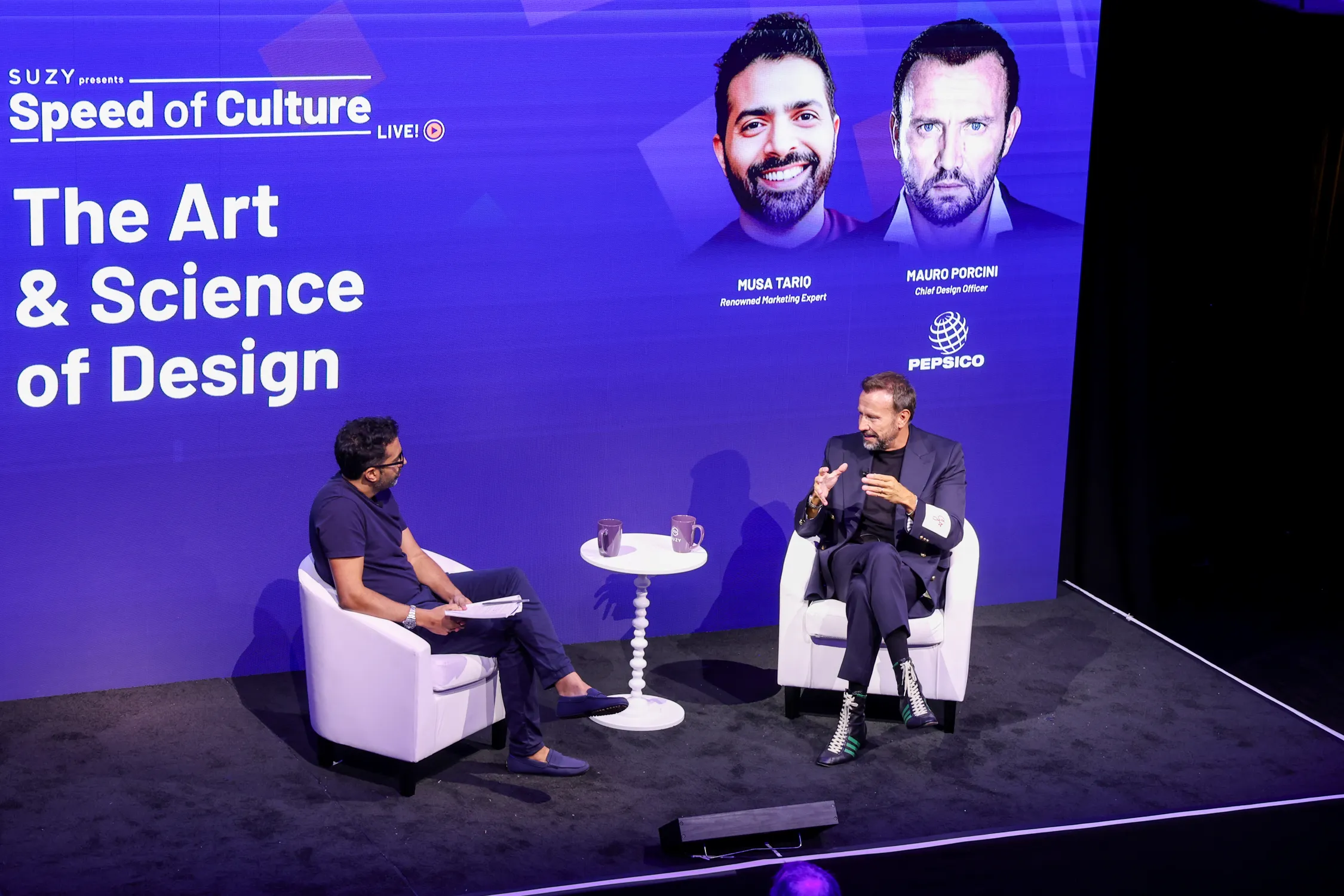 Two men seated on white armchairs on stage discussing design, with a large blue backdrop showing 'The Art & Science of Design' and portraits of Musa Tariq and Mauro Porcini from PepsiCo.