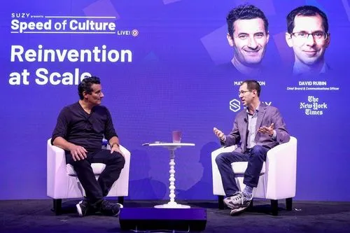 Two men seated on white armchairs on stage with a small white table between them, discussing in front of a purple backdrop reading 'Speed of Culture Reinvention at Scale' with headshots and names of Matthew and David Rubin.