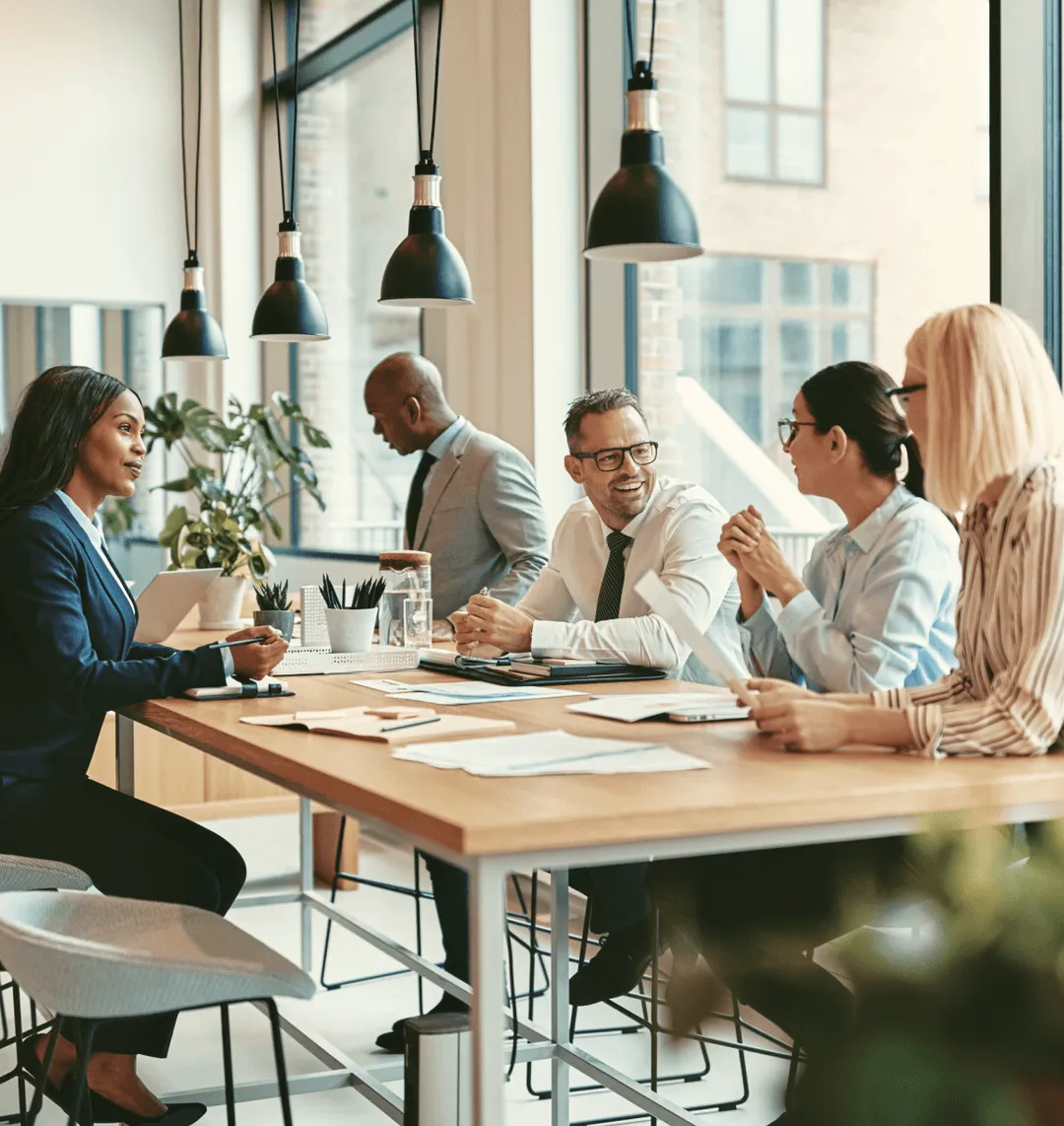 Five diverse business professionals in a modern office having a meeting around a wooden table with documents and plants.