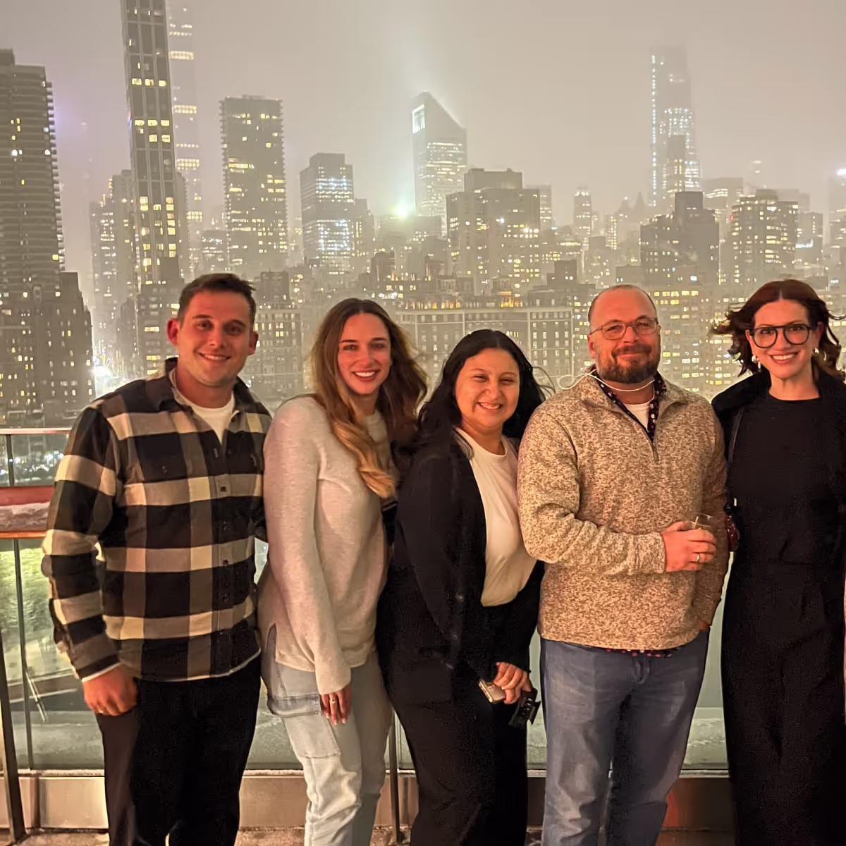 Five adults smiling and posing together on a rooftop at night with a city skyline illuminated in the background.