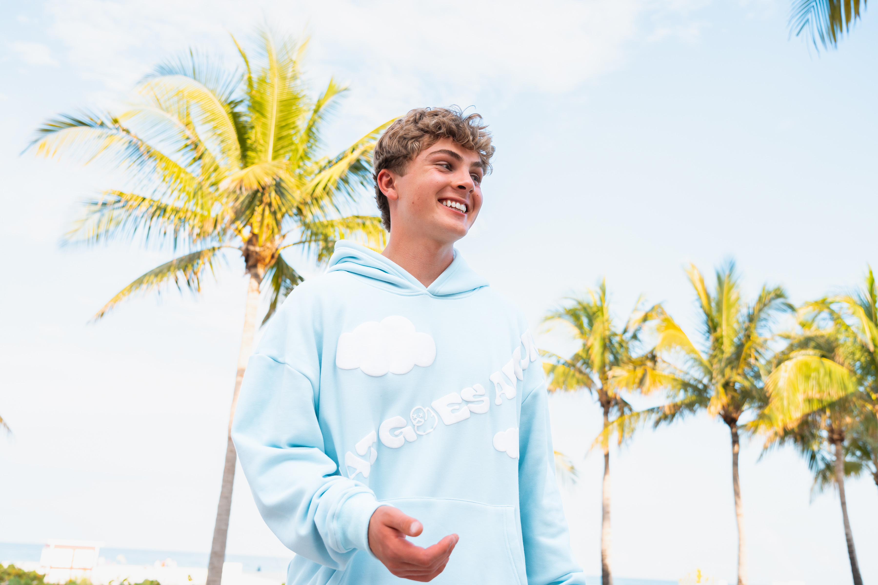 Young man smiling and wearing a light blue hoodie with cloud designs, standing outdoors with palm trees and a bright sky in the background.