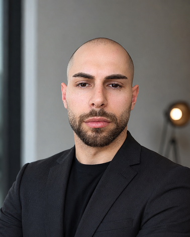 Portrait of a bald man with a beard wearing a black suit jacket and black shirt indoors.