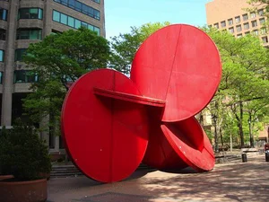 Red steel disc sculpture standing in a tree-lined plaza