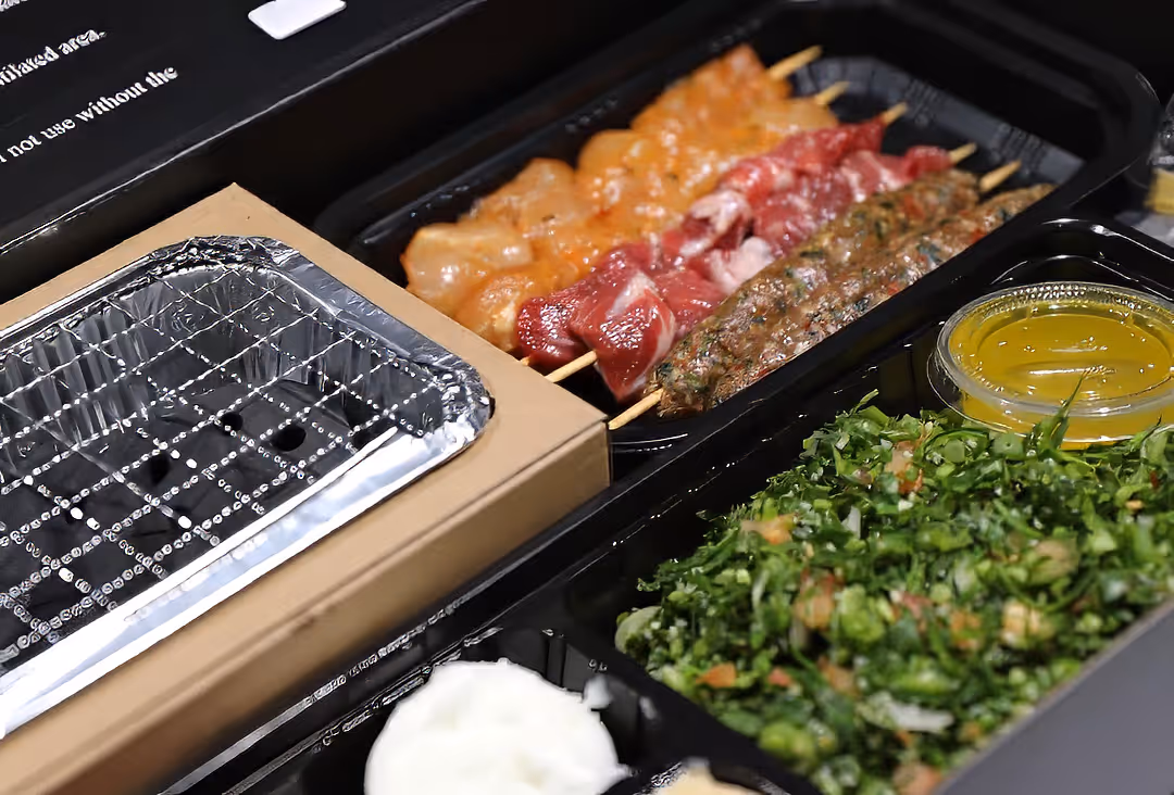Takeout meal box containing raw meat skewers, chopped green salad, yellow sauce, and a small portion of white sauce.