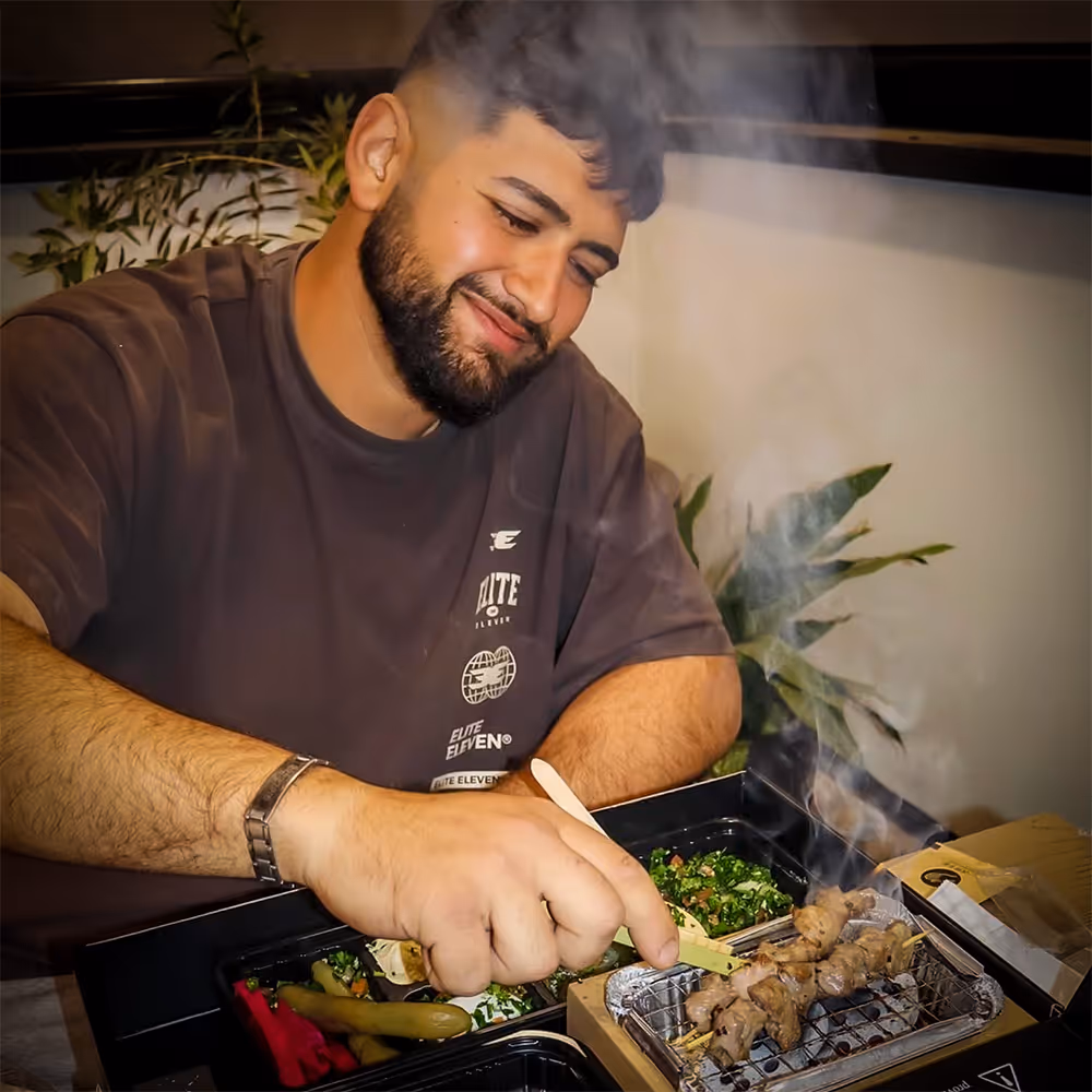 Smiling man grilling skewered meat on a small indoor grill with side dishes nearby.
