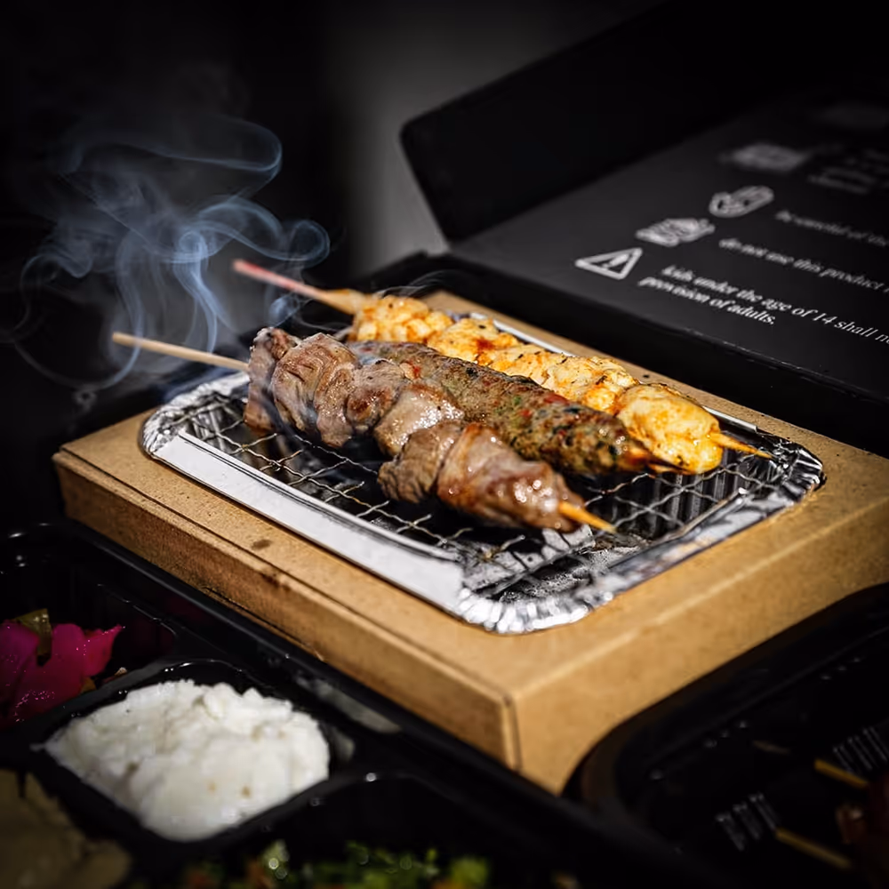 Three skewers of grilled meat and seafood cooking on a small portable tabletop grill with smoke rising.