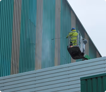 Person in yellow protective clothing and helmet using a lift to clean a tall green industrial building wall with a power washer.