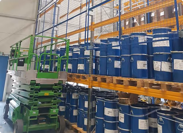 Grüner mobiler Scherenlift neben Regalen mit blauen Metallfässern in einem Lagerhaus.