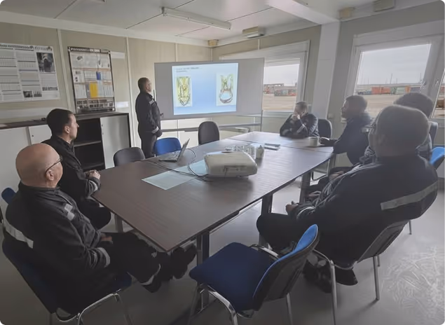 Männer in Arbeitskleidung sitzen an einem Konferenztisch und sehen einer Präsentation mit Pflanzenabbildungen auf einem Bildschirm zu.