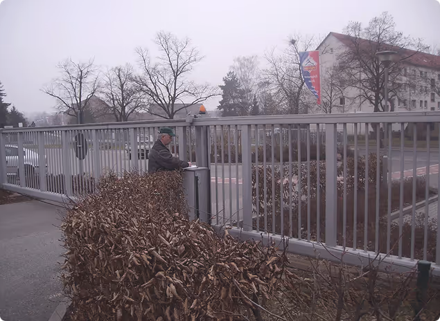 Ein älterer Mann steht an einem geschlossenen Metalltor neben einem Strauch mit braunen Blättern, im Hintergrund Häuser und kahle Bäume an einem bewölkten Tag.